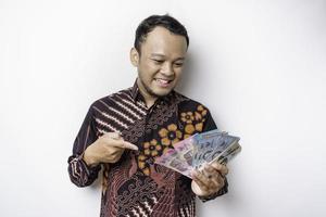 A happy young Asian man is wearing batik shirt and holding cash money in Indonesian rupiah isolated by white background photo