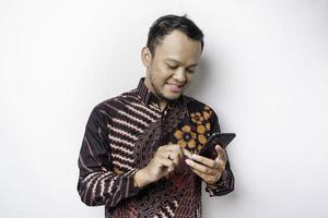 un retrato de un hombre asiático feliz con un pantalones batik y sosteniendo su teléfono, aislado de fondo blanco foto