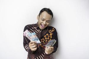 A happy young Asian man is wearing batik shirt and holding cash money in Indonesian rupiah isolated by white background photo