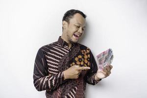 A happy young Asian man is wearing batik shirt and holding cash money in Indonesian rupiah isolated by white background photo