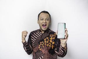 A happy young Asian man wearing batik shirt showing successful expression showing copy space on his phone isolated by white background photo