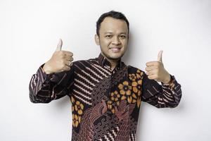 Excited Asian man wears batik shirt, gives thumbs up hand gesture of approval, isolated by white background photo
