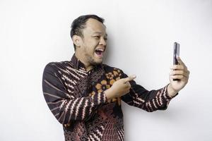 hombre asiático conmocionado con camisa batik y sosteniendo su teléfono, aislado de fondo blanco foto