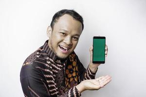 A portrait of a smiling Asian man wearing a batik shirt and showing green screen on her phone, isolated by white background photo