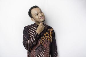 Thoughtful Asian man wearing batik shirt is looking aside while touching her chin, isolated by white background. photo