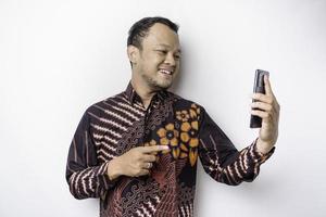 un retrato de un hombre asiático sonriente que lleva un pantalones de batik y muestra una pantalla verde en su teléfono, aislado de fondo blanco foto