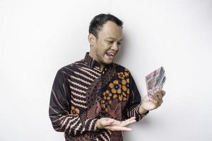 A happy young Asian man is wearing batik shirt and holding cash money in Indonesian rupiah isolated by white background photo