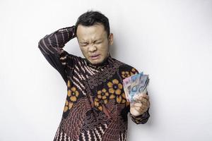 A stressed Asian man wearing batik shirt and holding money in Indonesian Rupiah isolated by white background looks depressed photo