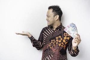 A happy young Asian man is wearing batik shirt, pointing at the copy space beside him while holding money in Indonesian rupiah isolated by white background photo