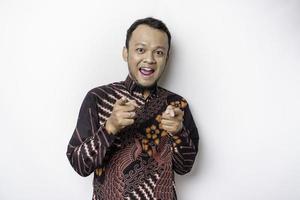 Young Asian man wearing batik shirt standing over isolated white background pointing fingers to camera with happy face. Good energy and vibes. photo