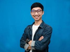 joven hombre asiático jeans camisa brazos cruzados sonrisa suave retrato fondo azul foto