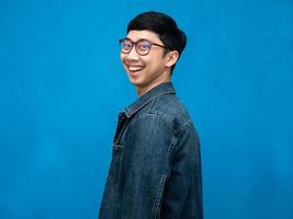 un hombre alegre usa anteojos camisa de jeans da la vuelta para mirar con una sonrisa feliz de fondo azul foto