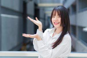 Professional young Asian working girl who wears white shirt is pointing hand to present something while  outdoors in the city with an office building in the background. photo