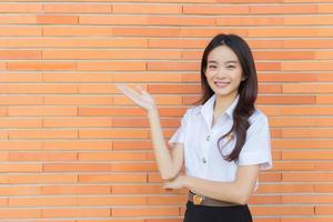 Portrait of an adult Thai student in university student uniform. Asian beautiful girl standing to present something confidently on brick walls background. photo