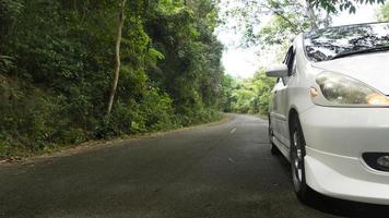 coche blanco conduciendo por la carretera asfaltada mojada. vista frontal del coche blanco con luz de señal de encendido. bosque verde al lado de la carretera. foto