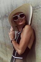 portrait of a woman in a swimsuit, hat and sunglasses in summer on the riverbank by a concrete wall photo