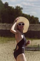 portrait of a woman in a swimsuit, hat and sunglasses in summer on the riverbank against the sky photo