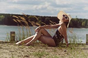 mujer en traje de baño, sombrero y gafas de sol toma el sol en verano en la orilla del río entre la hierba foto