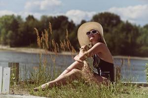 mujer en traje de baño, sombrero y gafas de sol toma el sol en verano en la orilla del río entre la hierba foto