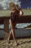 woman in a swimsuit, hat and sunglasses stands on the sand in summer on the riverbank at a concrete pier photo