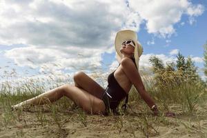 mujer en traje de baño, sombrero y gafas de sol toma el sol en verano en la orilla del río entre la hierba foto