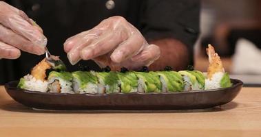 Küchenchef legt in Zeitlupe schwarzen Sushi-Kaviar auf Garnelen-Tempura-Sushi-Rollen mit Scheiben frischer Avocado - Nahaufnahme