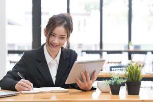 bella mujer de negocios asiática usando su laptop para trabajar y disfrutar trabajando, tomando notas, revisando tareas y sonriendo en la oficina. foto