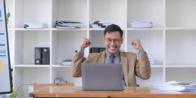 Happy young businessman in suit looking at laptop excited by good news online, lucky successful winner man standing at office desk raising hand in yes gesture celebrating business success win result photo