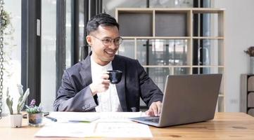 Young Asian businessman in suit and holding hot coffee cup and looking on laptop computer in office. Man in suit using laptop in well-lit workplace. photo