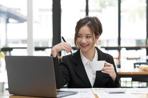 Beautiful Asian business woman using her laptop to work and enjoy working, taking note, reviewing assignment and smiling in the office. photo