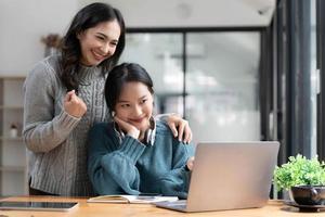 Two young Asian women show joyful expression of success at work smiling happily with a laptop computer in a modern office. photo