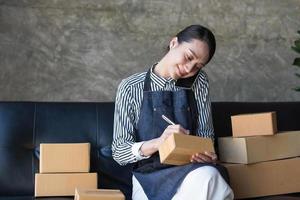 Empresaria de pequeñas empresas emergentes, pyme, mujer independiente que trabaja con un teléfono inteligente, joven propietaria de una pequeña empresa asiática que usa una computadora, entrega de cajas de embalaje en el mercado en línea, concepto de telemarketing de comercio electrónico de pymes foto