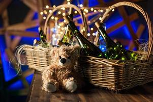 Teddy bear, champagne bottle with Christmas basket, gold garlands on dark brown wooden background, new year celebration concept photo