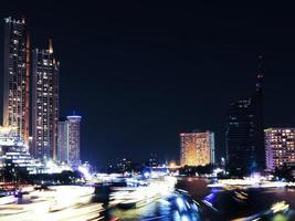 hermosas luces de transporte público acuático, tráfico de botes en el río chao phraya con edificios de condominios. hoteles de noche en bangkok, capital de tailandia foto