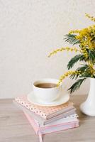 Bouquet of mimosa in a white vase, a cup of tea on a stack of notebooks for notes photo