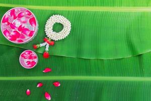 fondo del festival songkran con flores de guirnalda de jazmín en un tazón de agua, perfume y piedra caliza sobre un fondo verde de hoja de plátano húmedo. foto