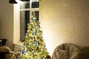 Christmas tree in white interior of a house with loft-style brick walls with garlands of glass toys on a rope. Glowing fairy lights decoration of the studio room. Cozy home in christmas and new year photo