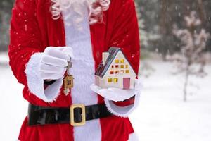 llave de la casa con llavero cabaña en manos de santa claus al aire libre en la nieve. trato de bienes raíces, compra, construcción, reubicación, hipoteca. hogar acogedor. feliz navidad, evento de reserva de año nuevo y sala foto