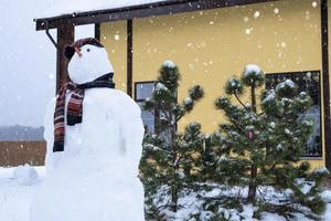 divertido muñeco de nieve con sombrero y bufanda en el fondo de una casa amarilla en el patio. invierno, entretenimiento de invierno, nevadas foto