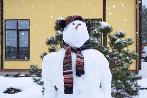 divertido muñeco de nieve con sombrero y bufanda en el fondo de una casa amarilla en el patio. invierno, entretenimiento de invierno, nevadas foto