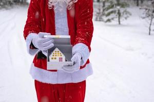 llave de la casa con llavero cabaña en manos de santa claus al aire libre en la nieve. trato de bienes raíces, compra, construcción, reubicación, hipoteca. hogar acogedor. feliz navidad, evento de reserva de año nuevo y sala foto