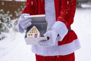 llave de la casa con llavero cabaña en manos de santa claus al aire libre en la nieve. trato de bienes raíces, compra, construcción, reubicación, hipoteca. hogar acogedor. feliz navidad, evento de reserva de año nuevo y sala foto