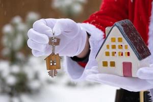 House key with keychain cottage in hands of Santa Claus outdoor in snow. Deal for real estate, purchase, construction, relocation, mortgage. Cozy home. Merry Christmas, new year booking event and hall photo