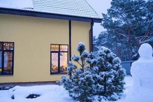 Cozy festive window of the house outside with the warm light of fairy lights garlands inside - celebrate Christmas and New Year in a warm home. Christmas tree, bokeh, snow on pine trees and snowfall photo