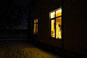 acogedora ventana festiva de la casa exterior con la cálida luz de las guirnaldas de luces de hadas en el interior: celebre la navidad y el año nuevo en un hogar cálido. árbol de navidad, bokeh, nieve en pinos y nevadas foto