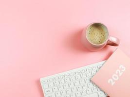 flat lay of computer keyboard, pink coffee cup  pink diary or planner  2023 on pink background with copy space. photo