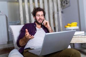 Young attractive smiling guy is browsing at his laptop and talking oh mobile phone, sitting at home on the cozy sofa, wearing casual outfit. Freelance business work from home concept photo