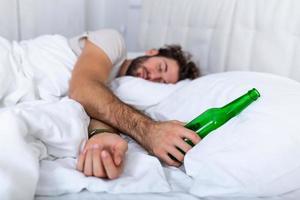 borracho en la cama y lugar triste y una botella de alcohol en la mano. hombre joven acostado en la cama mortalmente borracho sosteniendo una botella de alcohol casi vacía. foto