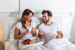 pareja enamorada desayunando en la cama. joven pareja caucásica desayunando romántico en la cama. hembra y macho, dos tazas de café, frutas y galletas coloridas. foto