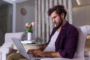 Man using laptop computer, working at home. Worried about finances. Handsome man with beard working at home on project, he is sitting on sofa looking at his laptop in front of him. Focus on the man photo